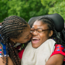 African American mother bends down to kiss her daughter on the cheek. The daughter is in a wheelchair