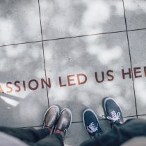 Two people standing on pavement. The perspective is looking down so you can see their shoes. "Passion Led Us Here" is printed on the pavement.
