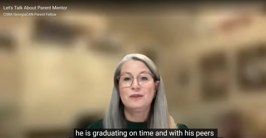 White woman with long grey straight hair and wire rim glasses is in a zoom field with a blurred background. Closed caption text says, he graduated with his peers