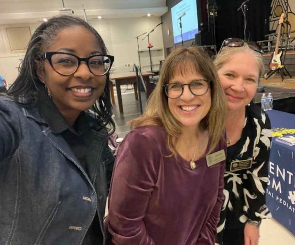 Three woman staning together for a photo. The woman on the left is a black woman with glasses and tight long braided hair. The woman in the middle is a white woman with glasses and dark blonde hair and the woman on the right is a white woman with light blond hair pulled back from her face