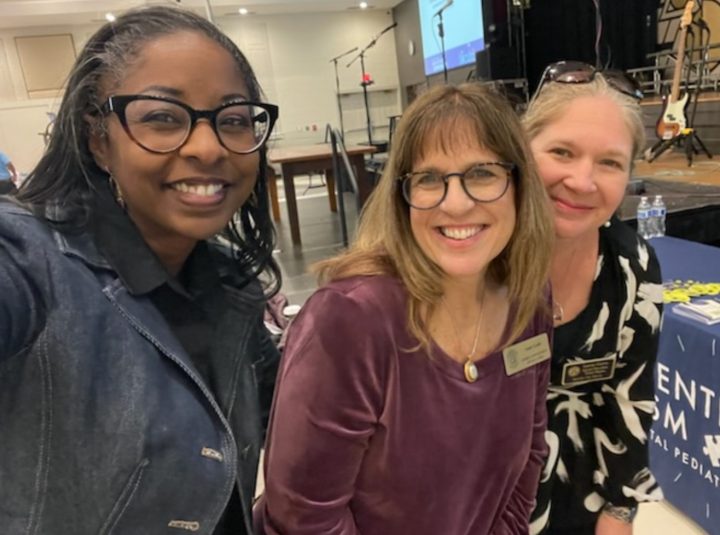 Three woman staning together for a photo. The woman on the left is a black woman with glasses and tight long braided hair. The woman in the middle is a white woman with glasses and dark blonde hair and the woman on the right is a white woman with light blond hair pulled back from her face