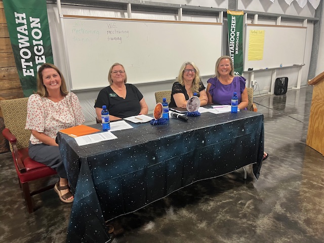 Four panelists sit at a table from the left are a white woman with dark shoulder length hair, a heavy set white woman with blond short hair and glasses, a white woman with blonde shoulder length wavy hair and glasses and a white woman with blonde curly hair